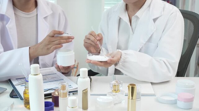 Two skincare professionals in white lab coats testing face cream at a desk with cosmetic bottles and jars, showing product development, beauty research, and clean modern work.