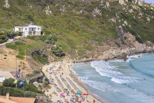 View of a sandy beach with rows of umbrellas and people, turquoise waves, and a lush green hillside with a white house in Santa Teresa Gallura, Sardinia, Italy.