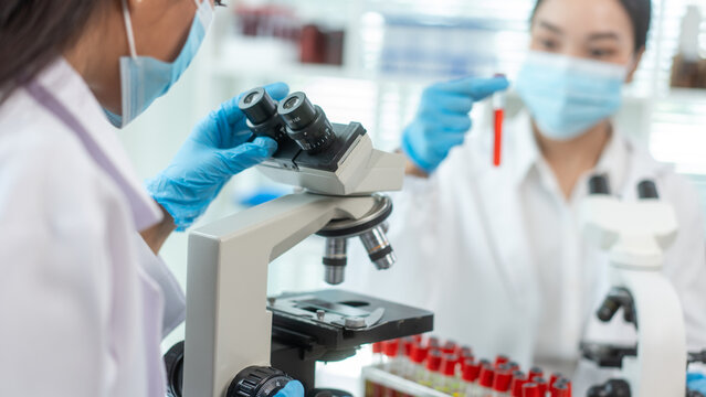 Scientists in medical masks and gloves work with microscopes and blood test tubes in a bright laboratory, showing teamwork, research, diagnosis, and healthcare science.