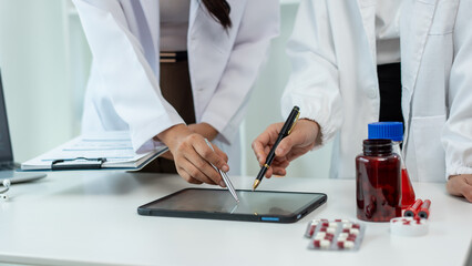 Obraz premium Medical professionals in white lab coats reviewing data on a tablet beside medicine bottles and pills on a clinic desk, showing teamwork, healthcare planning, and modern hospital work.