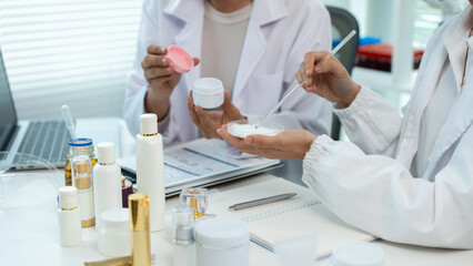 Two women in white lab coats test skincare samples at a bright office desk with bottles, laptop and notes, showing cosmetic research, teamwork and product development. © NanSan