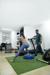 Fototapeta premium Female patient practicing balance and lower body stability on a BOSU trainer while being monitored by a physiotherapist during rehabilitation therapy.