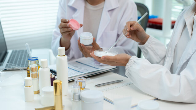 Two women in white lab coats test skincare samples at a bright office desk with bottles, laptop and notes, showing cosmetic research, teamwork and product development.