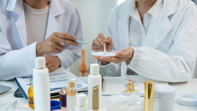 wo women in white lab coats test skincare samples at a bright office desk with bottles, laptop and notes, showing cosmetic research, teamwork and product development.