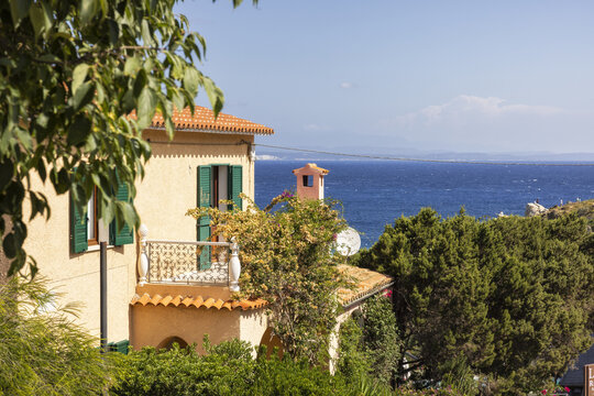 View of a Mediterranean house with orange walls and green shutters overlooking the blue sea and lush greenery in Santa Teresa Gallura, Sardinia, Italy.