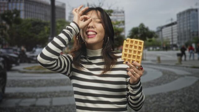 Woman holding waffle, fingers forming ok sign over eye, smiling with teeth visible on street with modern buildings and circular stone plaza; playful snack.
