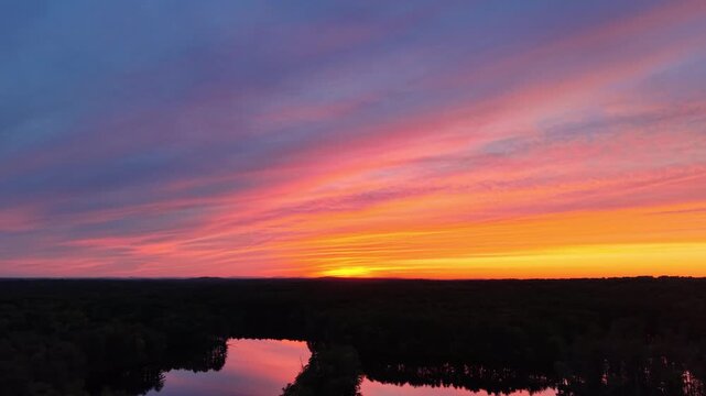 Drone view of colorful pink and orange sunset sky reflecting in calm lake surrounded by dark forest in Harold Parker State Forest, Massachusetts. Copy space