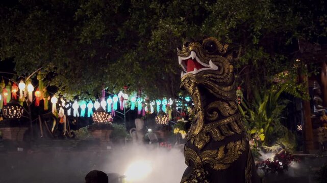 Static medium shot of an ornate golden Lanna-style Singha guardian lion statue with colorful festival lanterns and slowly drifting mist in foreground, mystical and cinematic night atmosphere