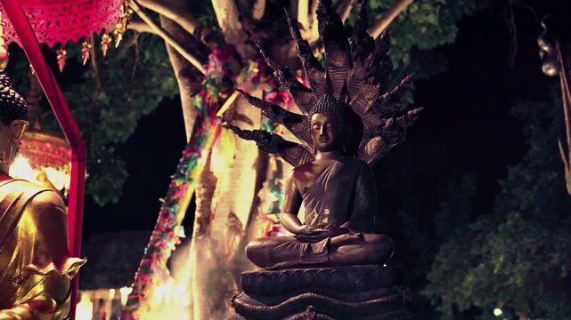Static medium shot of a Phra Nak Prok bronze Buddha in meditation pose sheltered by nine-headed Naga serpent with colorful Yi Peng garlands sacred Bodhi tree, and drifting mist at Northern Thai temple