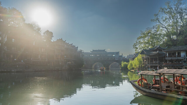 Wooden boats sail along the river. Canopies, lifebuoys on the decks. There are old traditional Chinese buildings on the shore. There is an arched stone bridge ahead. The sun in the blue sky. China.
 