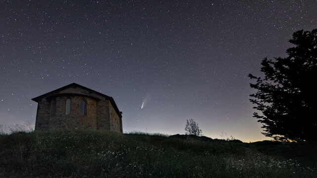 View of a small stone chapel on a grassy hill under a starry night sky featuring a bright comet with a visible tail and silhouetted trees.