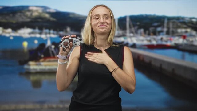 Woman holding optometry trial frame, hand on chest, smiling while wearing glasses at marina pier with moored boats and promenade; optical care gratitude.
