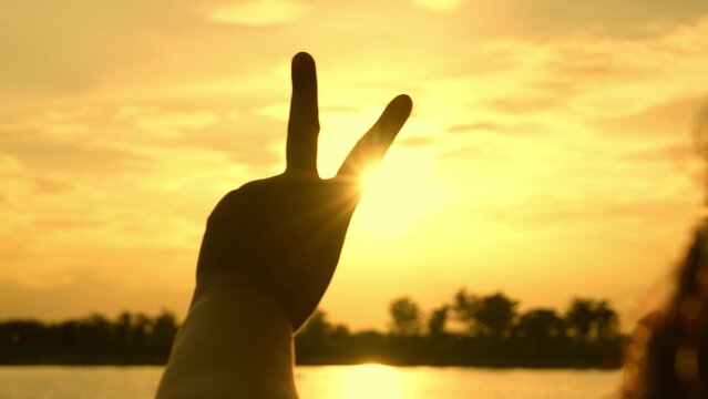 A hand forms the V-sign against a golden sky, evoking peace, victory, and dreams. The gesture also conveys strength, encouragement, progress, and a spirit of togetherness.
