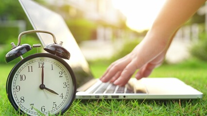 Time and technology meet as a clock stands before a laptop in a lush park, representing punctuality, self-discipline, mindful scheduling, eco-friendly workplaces, and smart digital routines.  © Punyawee