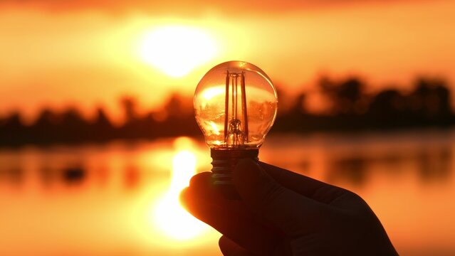 Holding a light bulb against a sunset lake backdrop symbolizes innovation, creative energy, inspiration from nature, the power of ideas, and the fusion of technology and the environment.
