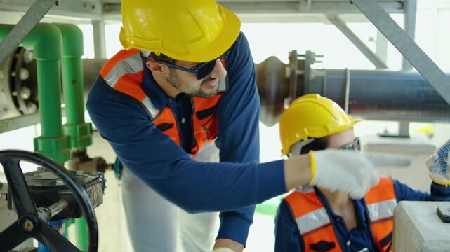 Two professional industrial engineers wearing protective safety gear carefully inspecting pipeline valves and discussing maintenance data on a digital tablet inside a large manufacturing plant.
