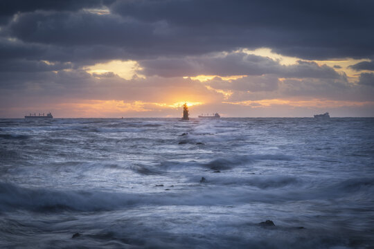 View of a lighthouse beacon amidst rough sea waves and cargo ships on the horizon during a dramatic sunset in Dublin, County Dublin, Ireland.