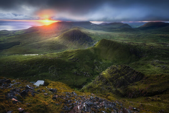View of the Coumanare mountains under a dramatic sunset sky with clouds over green slopes and rocky terrain in County Kerry, Ireland.
