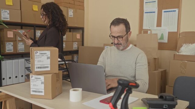 Man typing on laptop amid stacked shipping boxes and labels while woman holds clipboard taking notes in a building; focused teamwork.