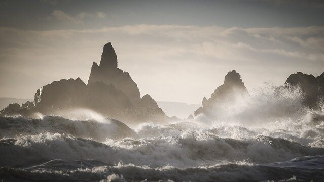 View of Kilfarrasy Beach featuring dramatic sea stacks and powerful white waves crashing against the rugged coastline under a misty sky in County Waterford, Ireland.