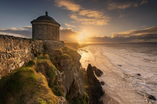 View of Mussenden Temple perched on a rugged cliff edge overlooking Downhill Strand beach during a golden sunset with dramatic clouds and waves Coleraine, Northern Ireland, United Kingdom.