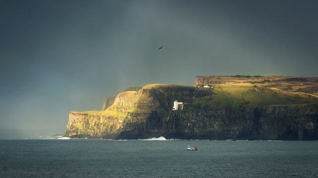 View of the dramatic cliffs of Rathlin Island featuring the West Light lighthouse and a small boat on the sea under a moody sky Rathlin Island, Northern Ireland, United Kingdom.