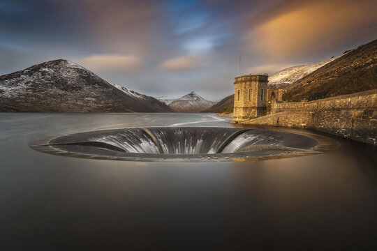 View of the bell-mouth spillway and stone tower at Silent Valley Reservoir with snow-dusted mountains under a sunset sky in Newry, Northern Ireland, United Kingdom.