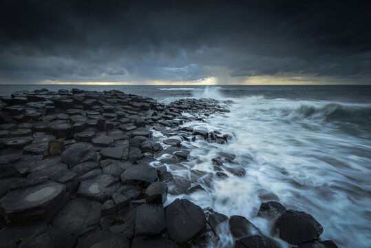 View of the hexagonal basalt columns of Giant's Causeway with waves crashing against the rocks under a dark stormy sky in Causeway Coast and Glens, Northern Ireland, United Kingdom.