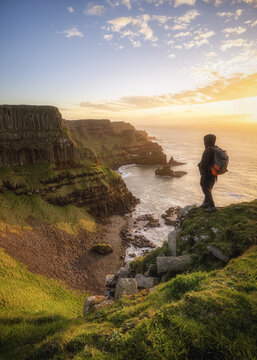 View of a hiker standing on a grassy cliff overlooking the basalt columns of Giant's Causeway at sunset in Causeway Coast and Glens, Northern Ireland, United Kingdom.