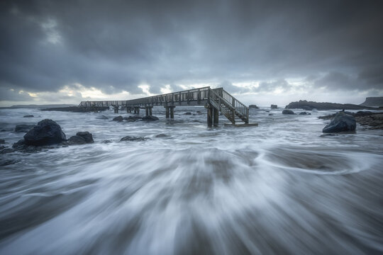 View of a wooden pier over the rushing sea with dark moody clouds and rocky shoreline in Ballintoy, Northern Ireland, United Kingdom.