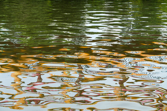 Rippling water with highly distorted colorful reflections of trees and structures. Fragments of white, golden-yellow, blue, subtle pinks and reds create fluid abstract patterns. Dynamic yet meditative