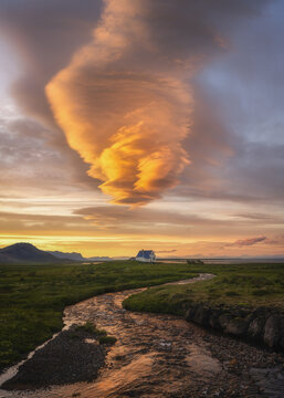 View of a small white house with a blue roof nestled in green fields by a winding stream under a dramatic glowing lenticular cloud at sunset Arnarstapi, Snaefellsbaer, Iceland.
