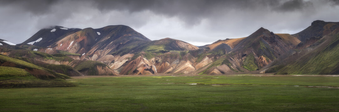 View of the colorful rhyolite mountains of Landmannalaugar with a lush green valley under a dramatic cloudy sky in Rangarthing ytra, Iceland.