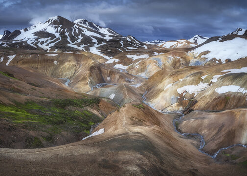 View of the colorful rhyolite mountains of Kerlingarfjoll with snow patches and a winding stream under a dramatic sky in Hrunamannahreppur, Iceland.