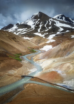View of the Kerlingarfjoll mountain range with a winding river, snow patches on rhyolite hills, and a small bridge under a dramatic sky in Hrunamannahreppur, Iceland.