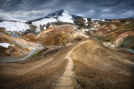 View of the Kerlingarfjoll mountains with a wooden staircase trail, snow patches, and orange rhyolite hills under a dramatic cloudy sky Hrunamannahreppur, Iceland.