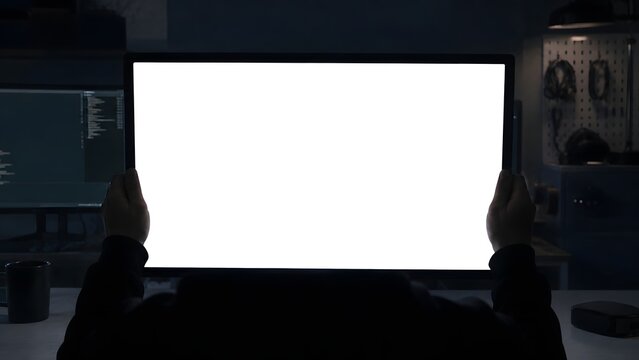 Person holding a large computer monitor with a bright white blank screen while sitting in a dark workspace featuring soft lighting and a professional office environment