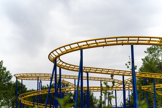 An outdoor shot of a winding yellow metal roller coaster track supported by blue pillars, surrounded by green trees under an overcast gray sky. 