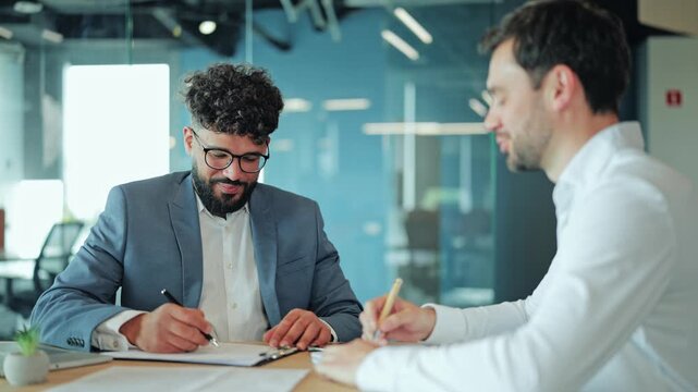 Two diverse businessmen sitting at desk during legal agreement. Middle Eastern man in suit smiling and looking at European colleague or lawyer who is writing on paper or contract document.