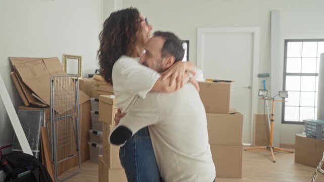 Man lifts woman in a tight hug among stacked moving boxes inside a building with visible worklight and hardhat on a box; joy new home together.