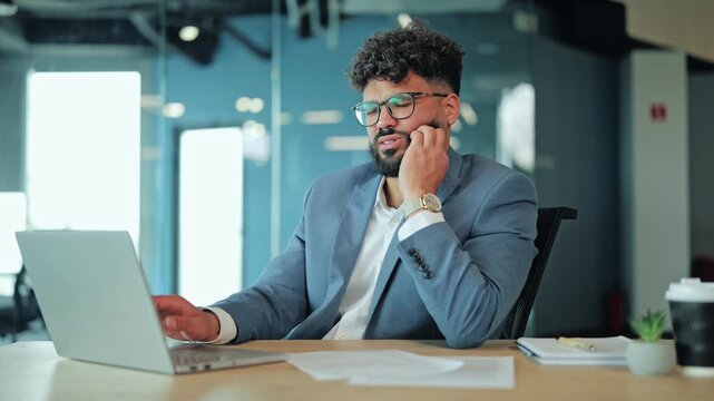 Middle Eastern businessman in a suit looking pained while holding his jaw due to a severe toothache. Image depicting dental health problems, discomfort, and stress during a workday at the office.
