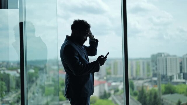 Corporate man silhouette standing at office window looking distressed. Person holds smartphone while reading bad news, face shows deep worry frustration about professional failure at workplace now.