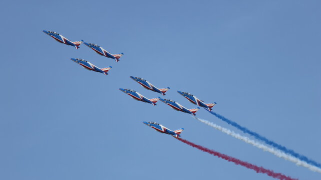 View of the Patrouille de France aerobatic team flying in formation with red, white, and blue smoke trails against a clear blue sky in La Ferte-Alais, Ile-de-France, France.