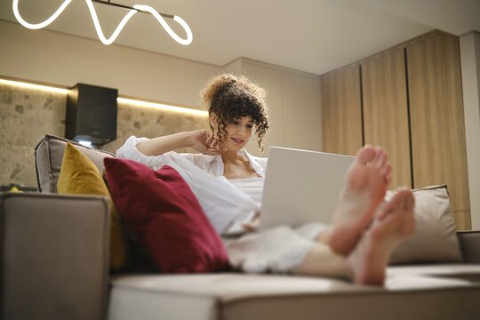 A young woman sits comfortably on a couch with her feet up, using her laptop for online video call in a bright living room decorated with warm, neutral tones. The atmosphere is inviting and relaxed