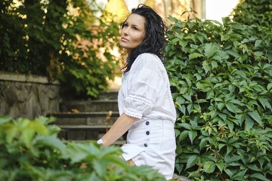 A woman with curly hair is seated gracefully on stone steps surrounded by vibrant green plants. She wears a stylish outfit and gazes thoughtfully into the distance on a sunny day