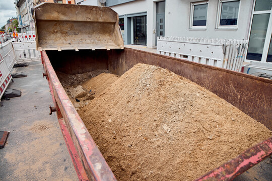 A large metal skip container filled with a pile of construction sand and soil next to an excavator bucket on a city street site.