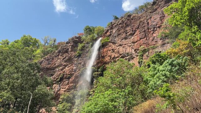 A tall, narrow waterfall tumbles dramatically down a rugged red-brown rocky cliff face surrounded by lush green tropical forest trees under a bright blue sky with scattered white clouds.