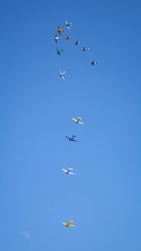 View of a formation of vintage propeller airplanes flying across a clear blue sky with a faint moon visible below La Ferte-Alais, Ile-de-France, France.