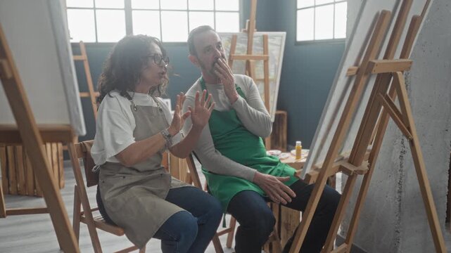 Woman gestures with open hand toward canvas while man in green apron watches and smiles, both seated by easels and paint bottles in an art studio; creative collaboration joy.