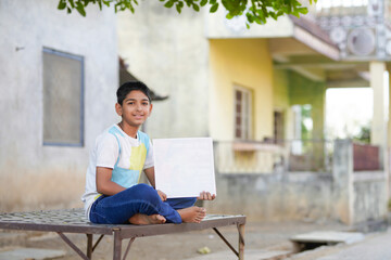 Fototapeta premium Indian schoolboy sitting outdoors in a rural village while holding and pointing at a blank display board